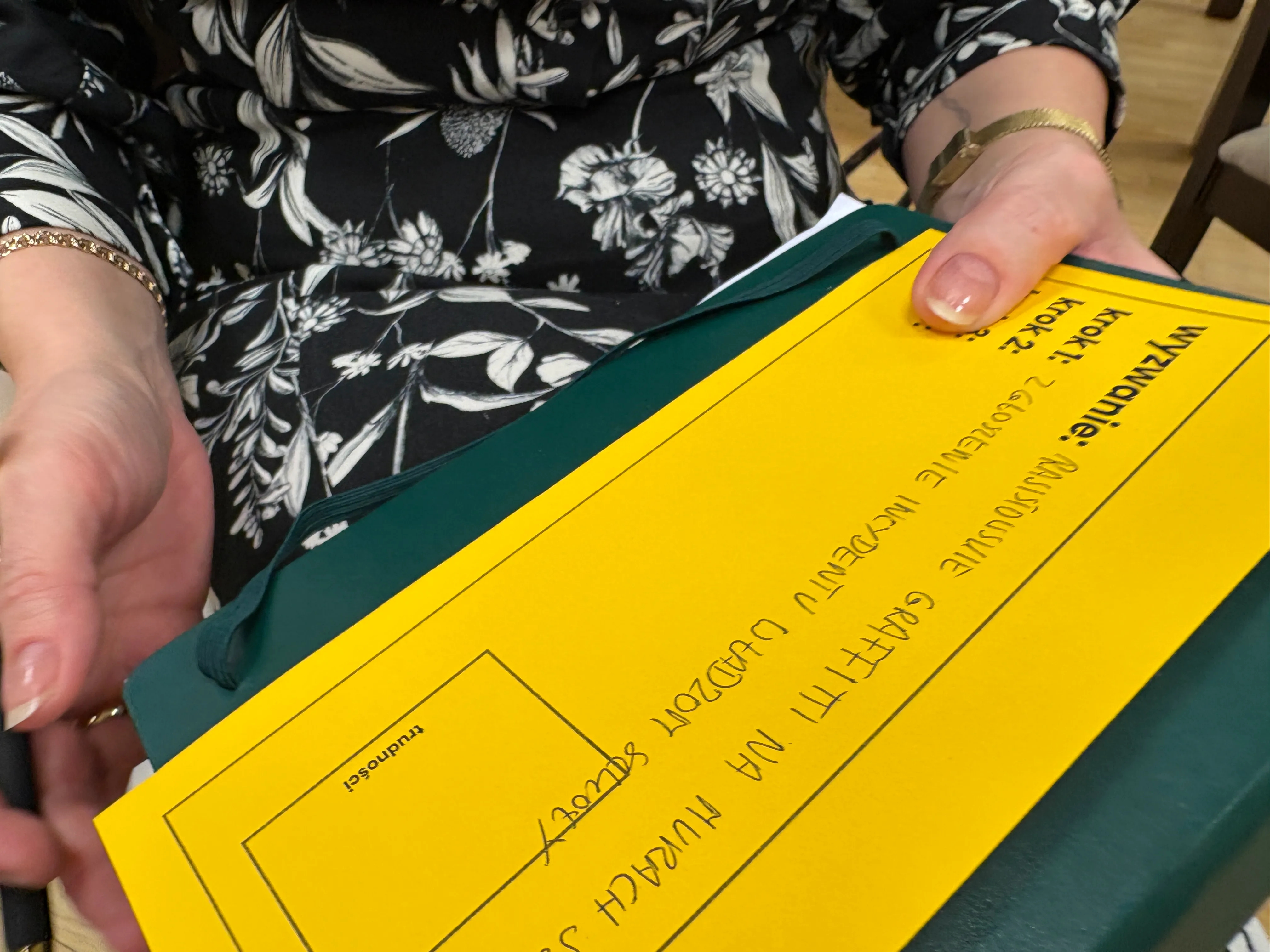 Dwie ręce trzymają zieloną teczkę z żółtą kartką papieru. Osoba ubrana jest w czarną bluzkę w biały kwiatowy wzór. Na nadgarstku lewej ręki znajduje się złota bransoletka.