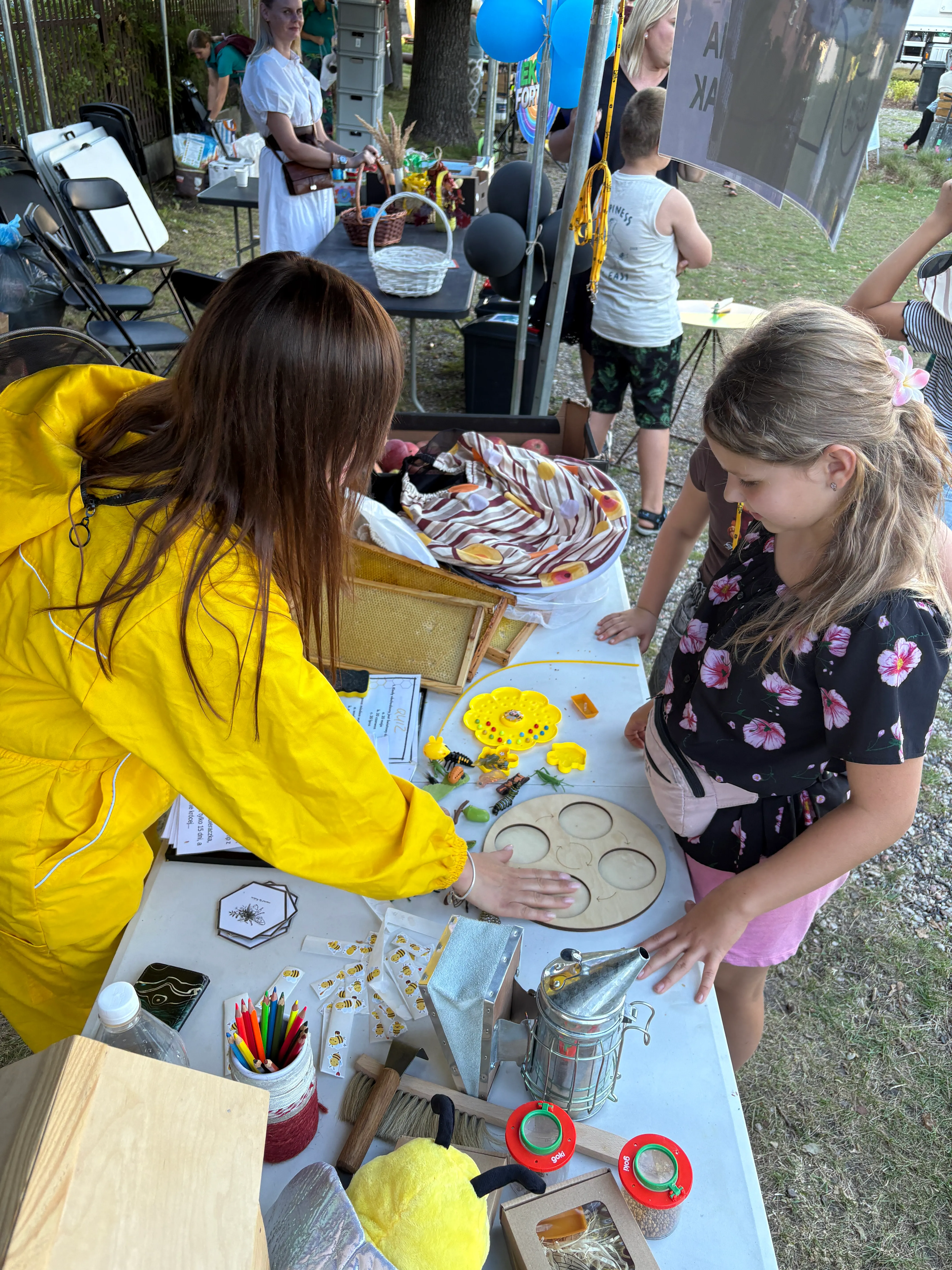Dwie dziewczynki stoją przy stoisku edukacyjnym na zewnątrz, gdzie na stole leżą różne przedmioty związane z pszczelarstwem i dzieci bawią się drewnianą układanką.