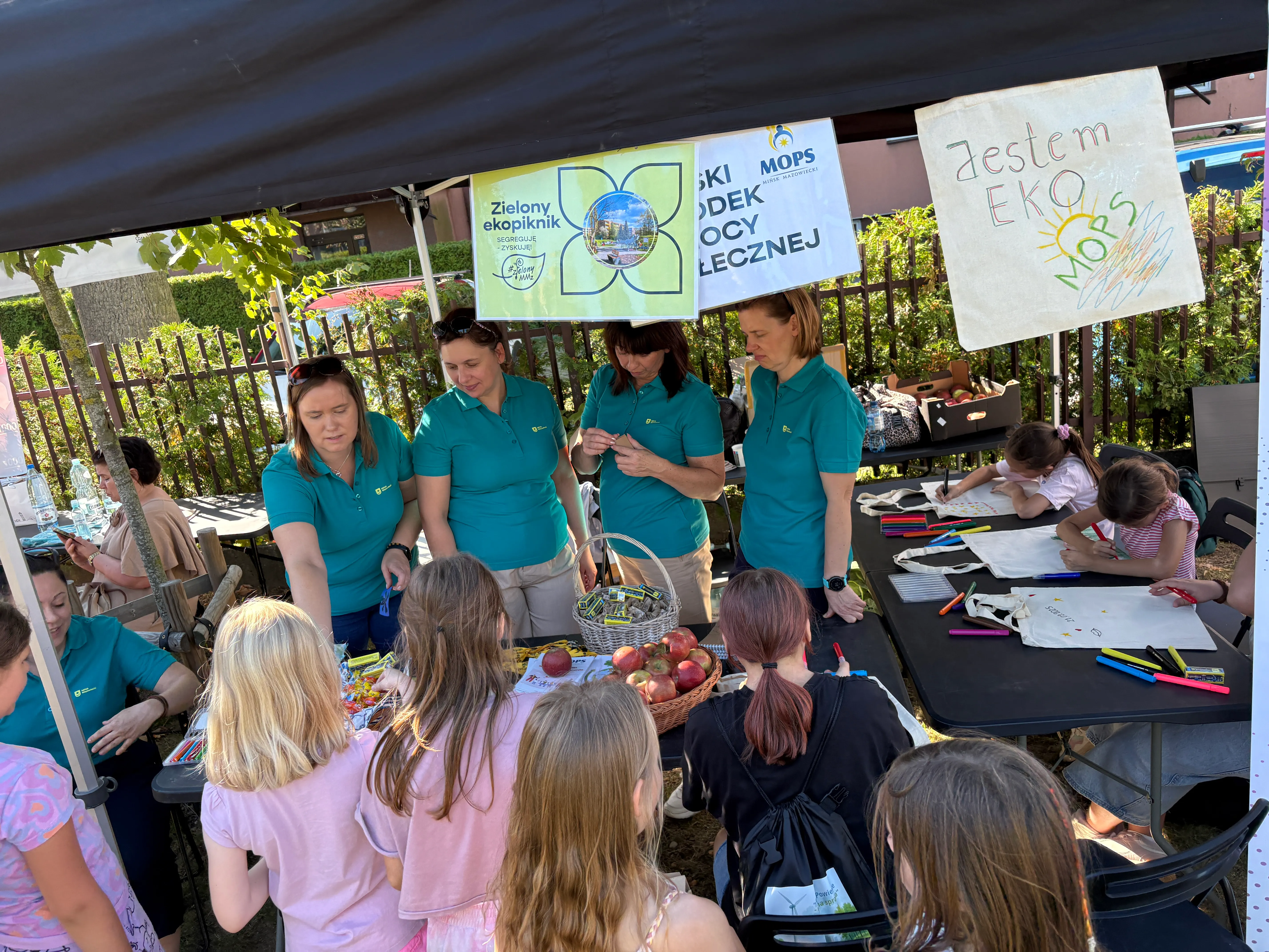 Pod namiotem na zewnątrz cztery kobiety w turkusowych koszulkach obsługują stoisko z jabłkami i materiałami edukacyjnymi, przed nimi stoi grupa dzieci, obok przy innym stole dwoje dzieci rysuje na materiałowych torbach.