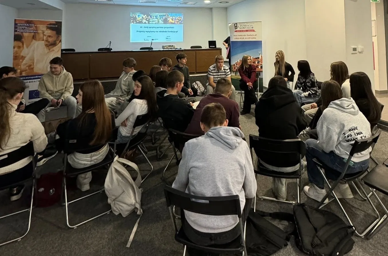 Grupa około dwudziestu młodych osób siedzi na krzesłach ustawionych w dwóch kołach w sali konferencyjnej z jasnymi ścianami i dywanem na podłodze. Na ścianie w tle znajduje się ekran z wyświetlanym tekstem oraz dwa stojące banery. Uczestnicy rozmawiają ze sobą, większość ma casualowy strój, niektórzy z nich mają plecaki obok krzeseł.